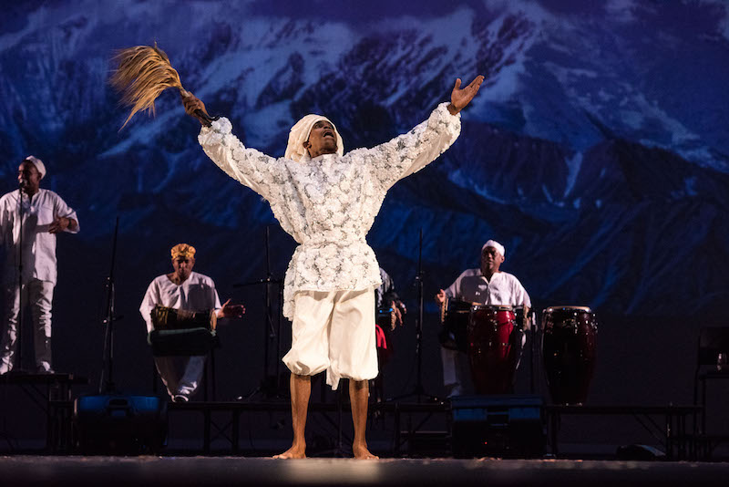 A man in white out stretches his arms while singing. Musicians are in the background.